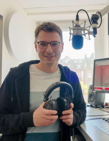 Mann mit Brille hält Kopfhörer in einem Studio mit Mikrofon und Bildschirm im Hintergrund. Tageslicht strömt durch ein Fenste